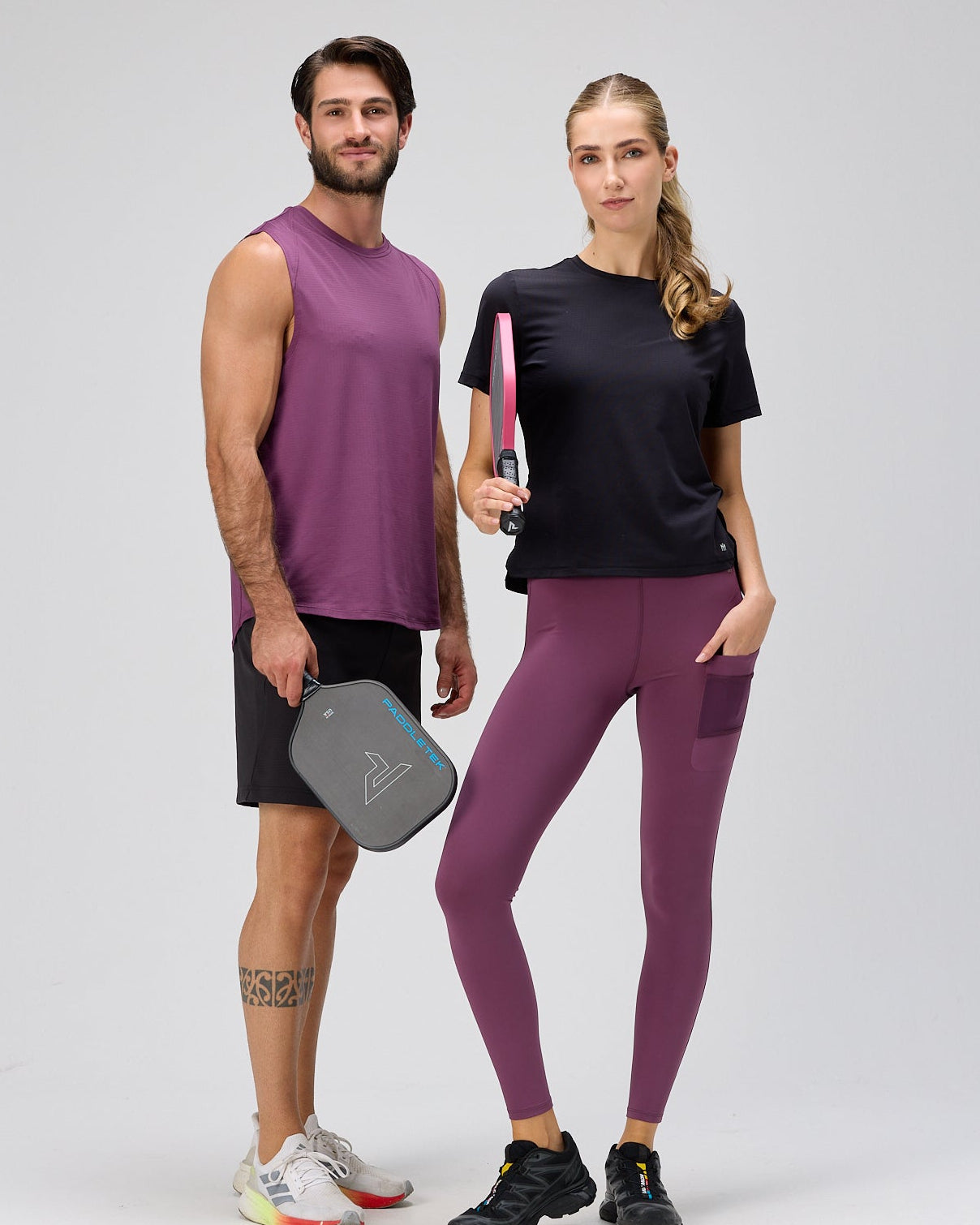 A man and a woman standing side-by-side in athletic wear against a light gray studio background. The man wears a purple sleeveless athletic shirt, black shorts, and white running shoes while holding a gray pickleball paddle.