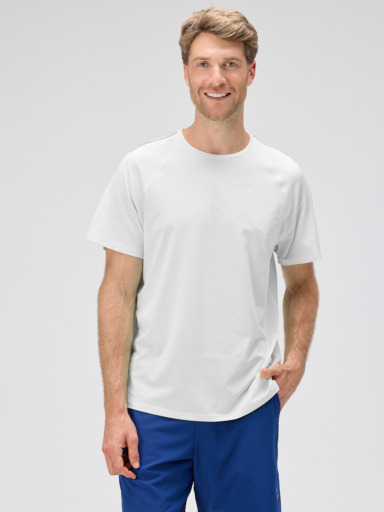 Man wearing a white lightweight moisture-wicking stretch jersey shirt with horizontal micro-perforations and bonded seams, paired with blue shorts for padel and pickleball.