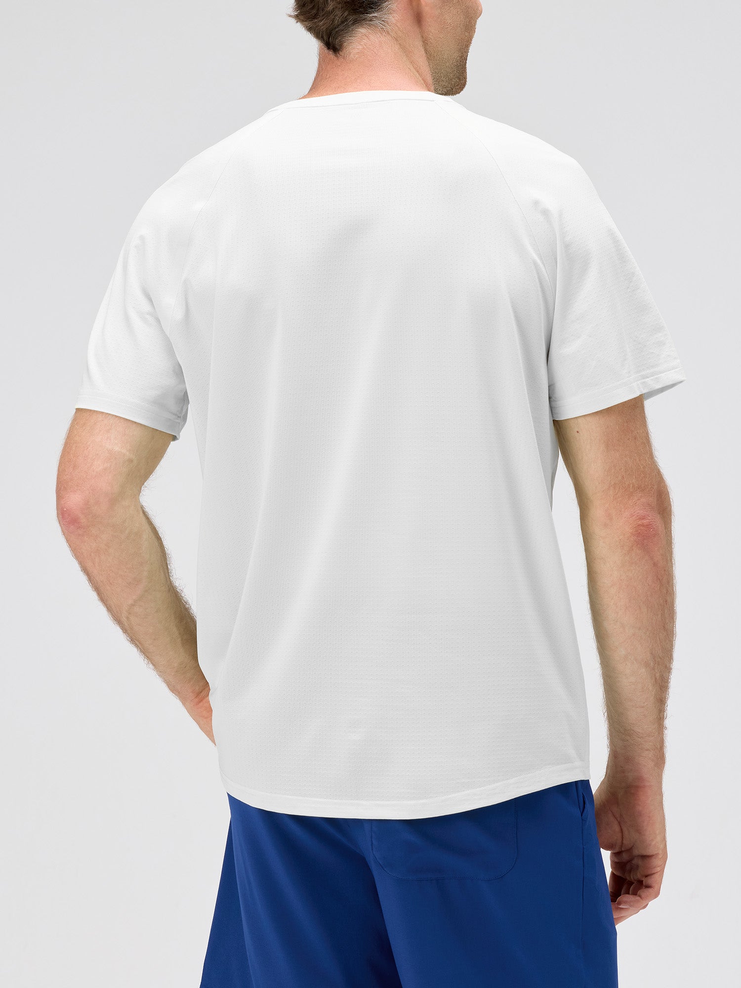Back view of a man wearing a white lightweight moisture-wicking stretch jersey with horizontal micro-perforations for airflow, paired with blue shorts for padel and pickleball sportswear.
