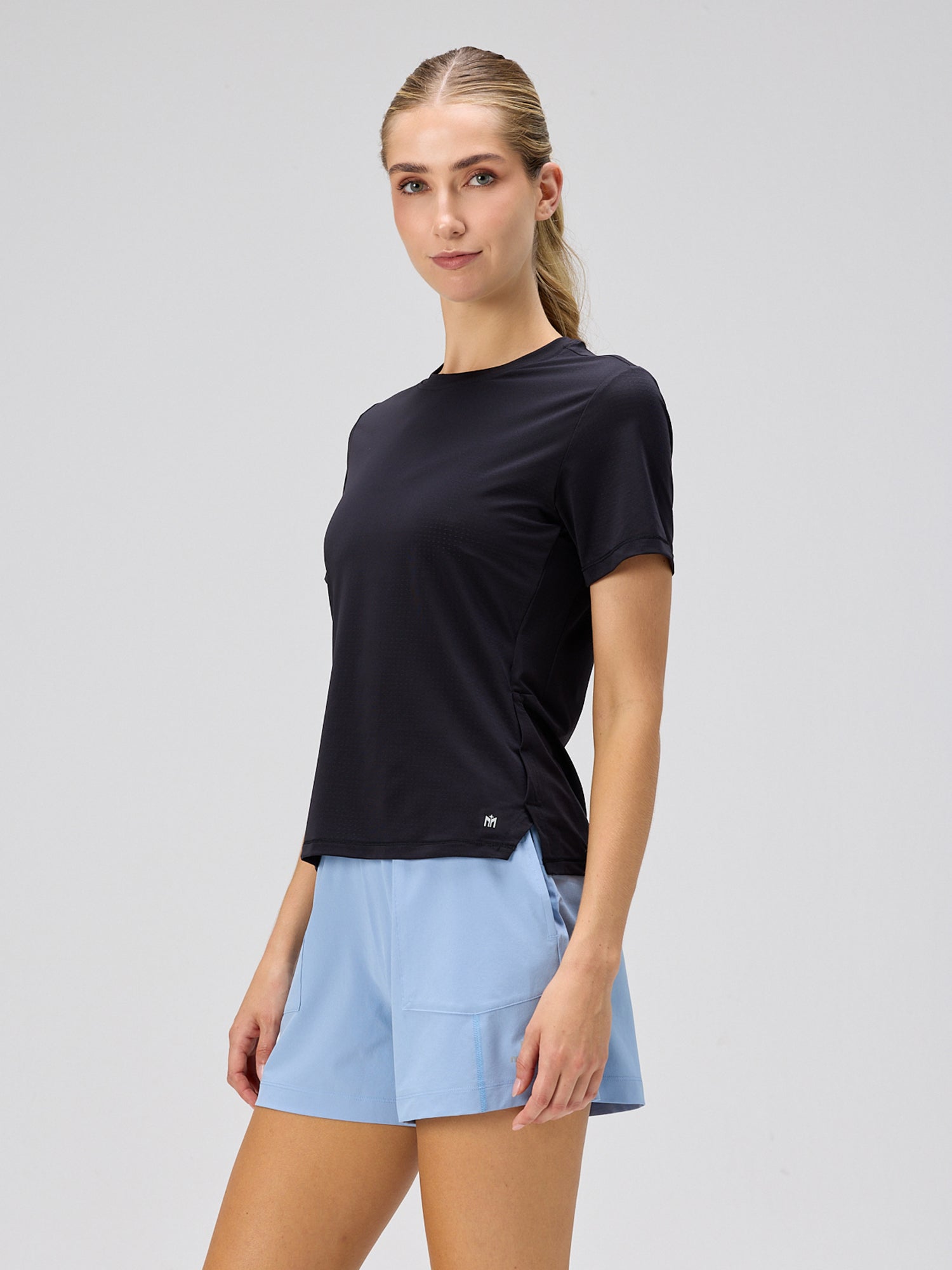 Woman wearing a black short-sleeve athletic shirt paired with light blue sporty shorts, standing against a plain light gray background.