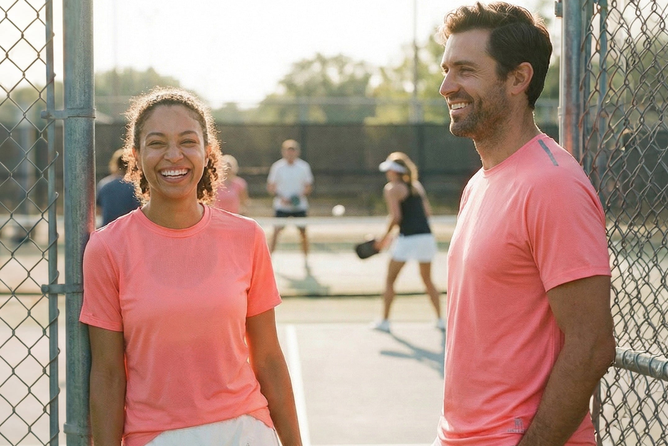 Two people in pink shirts standing on a tennis court with a chain-link fence in the background.