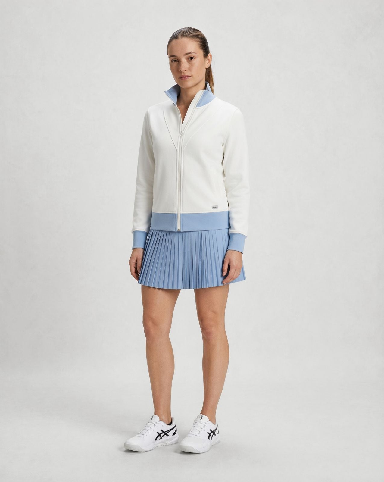 woman wearing white zip-up jacket with light blue trim and pleated light blue pickleball skort in minimal studio setting