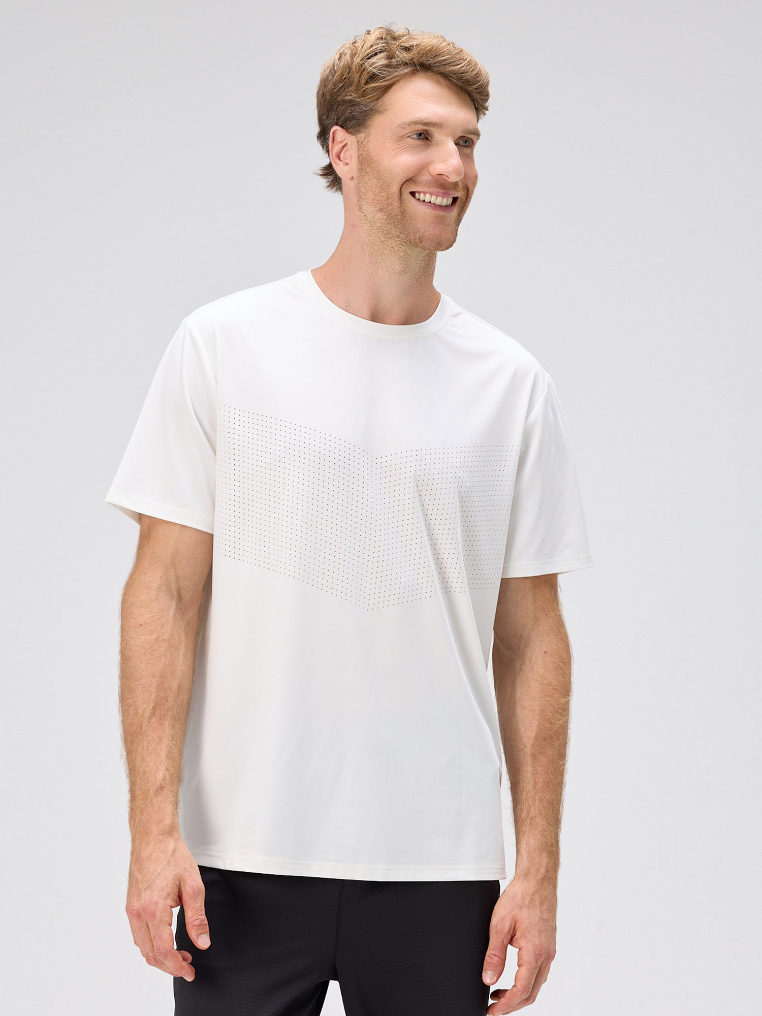 Man smiling and wearing a white short-sleeve tee with a subtle chevron pattern across the chest, paired with black shorts.