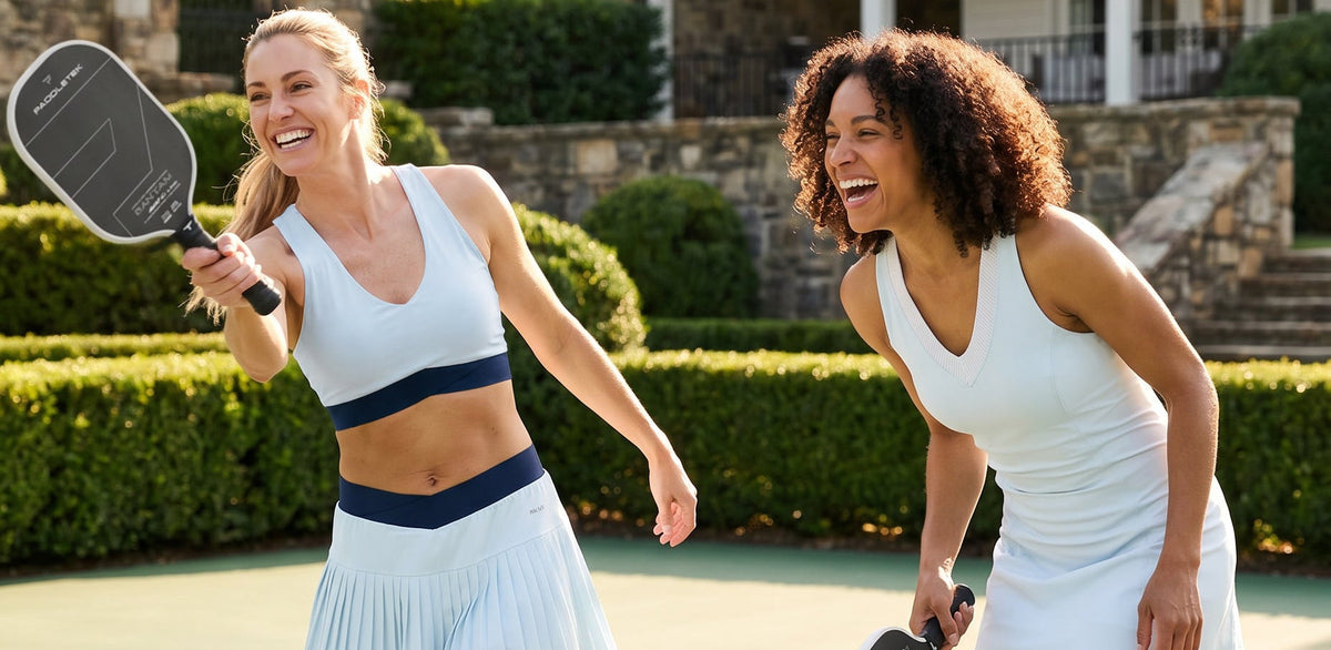 Two women laughing on an outdoor pickleball court, wearing matching white athletic dresses with navy blue waistbands and holding paddle-shaped exercise rackets.