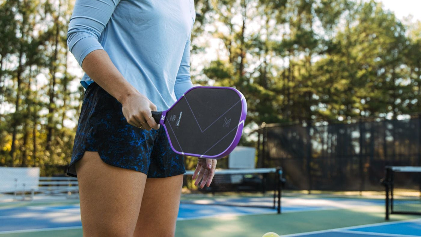 Woman holding a pickleball paddle on a court with trees in the background