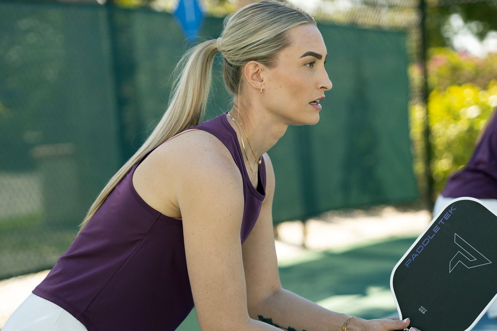 Two people playing pickleball on an outdoor court with a green fence and trees in the background.