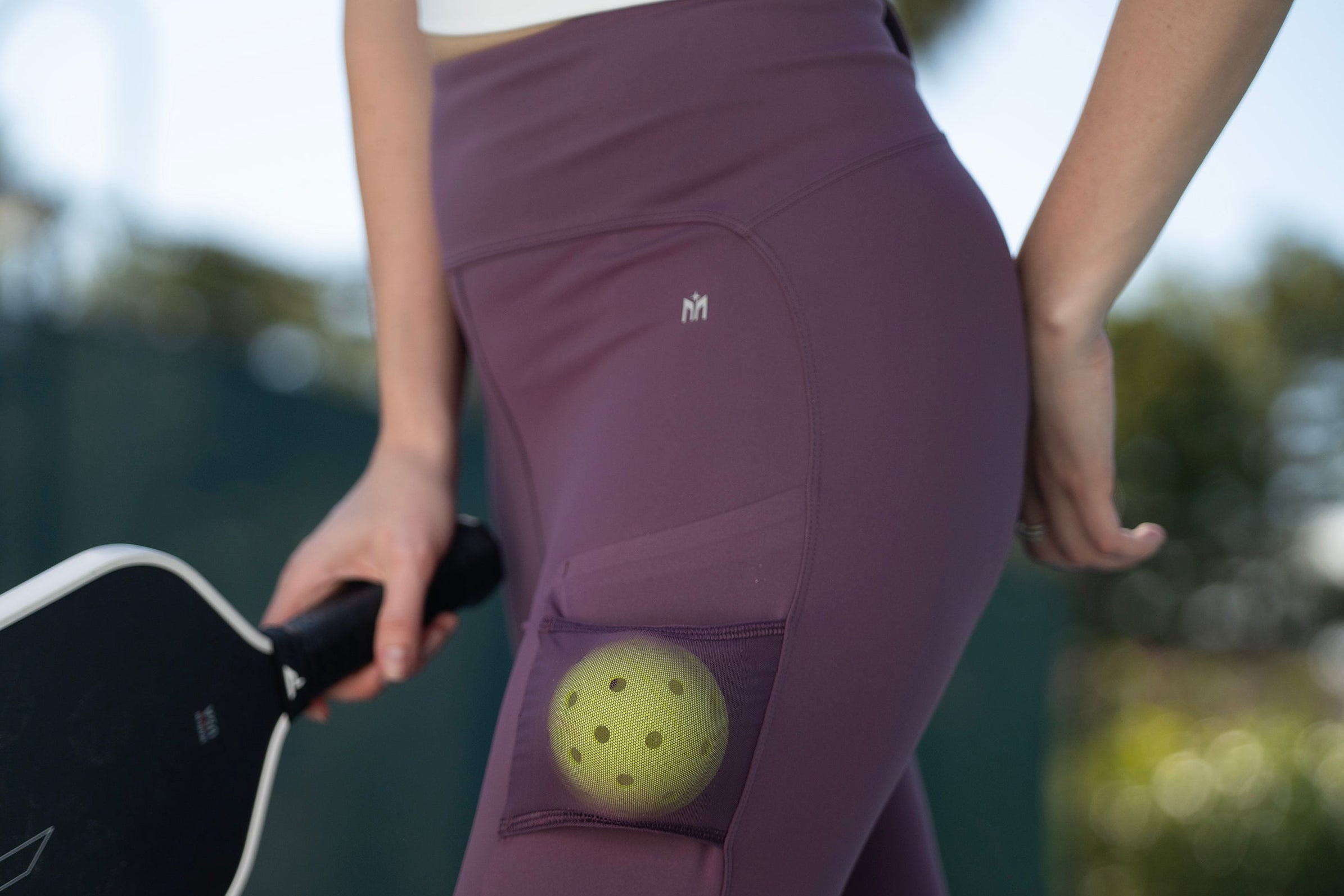 Person holding a pickleball paddle with a ball in a pocket of purple leggings on a blurred outdoor background