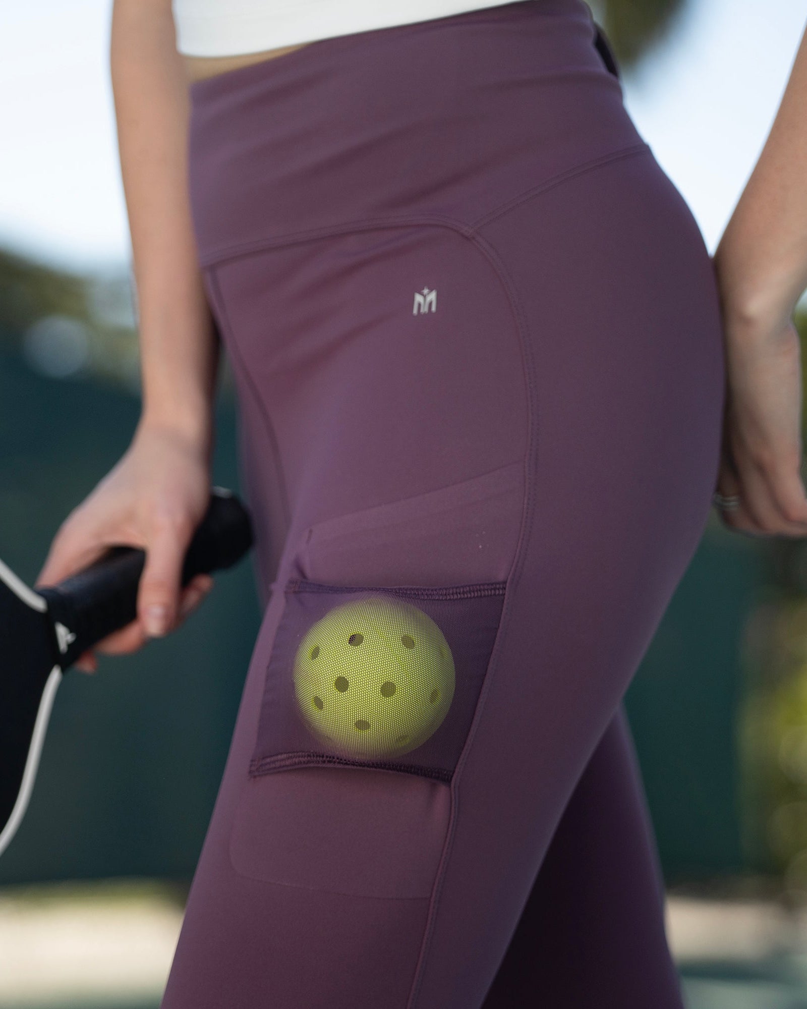 close-up of plum court leggings with mesh ball pocket holding a yellow pickleball during outdoor play