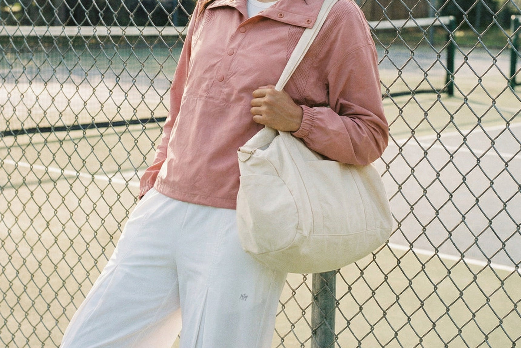 Woman in pink jacket and white pants standing on a tennis court with a fence in the background