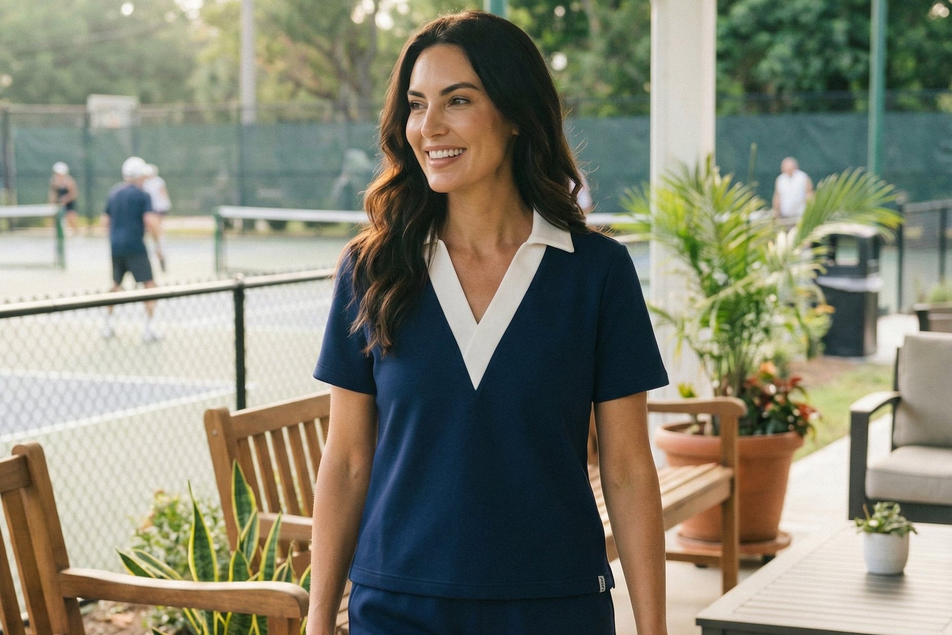 Woman in navy polo shirt and shorts standing on a patio with outdoor furniture and plants.