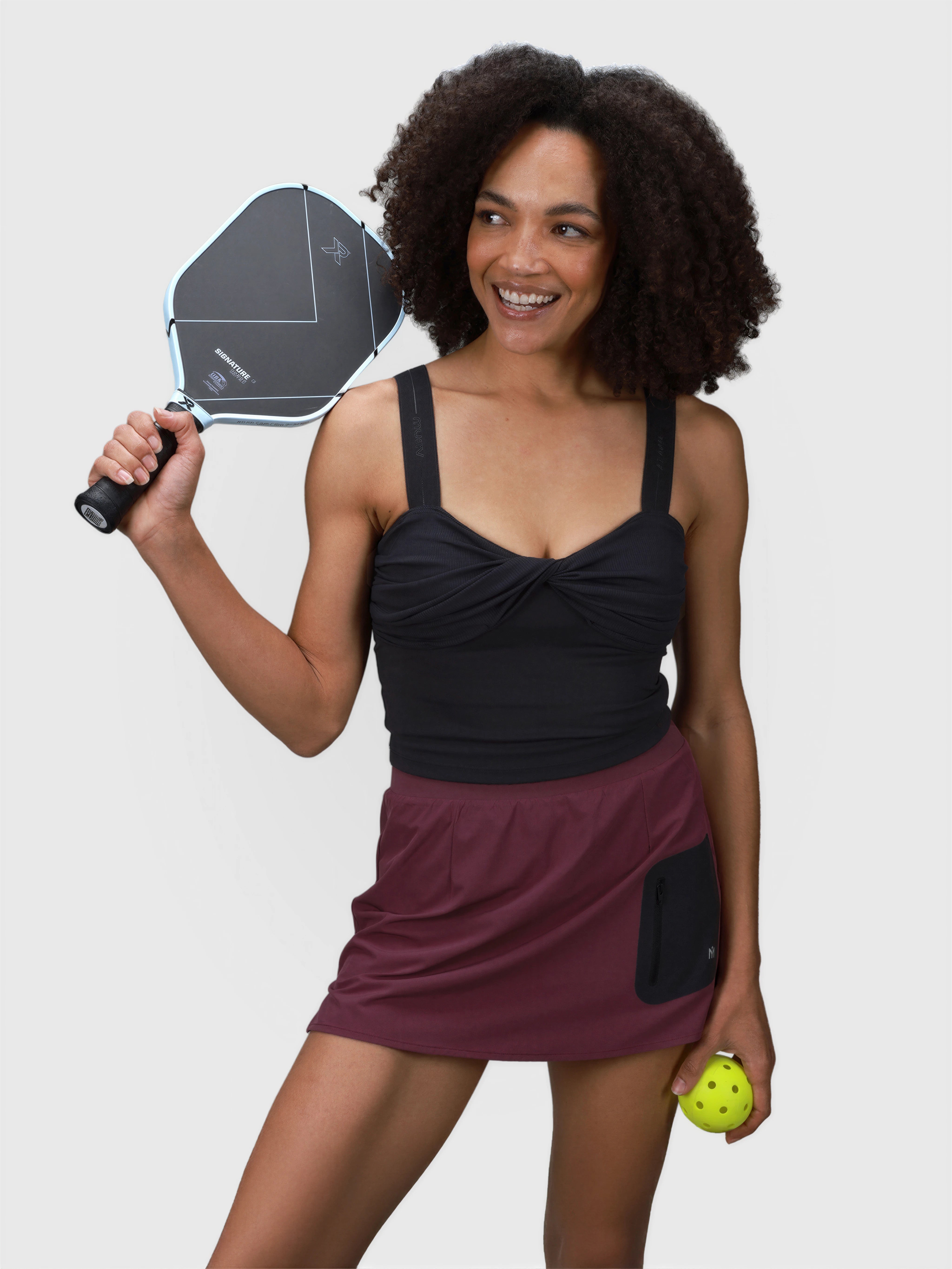 Woman wearing plum Motion Compression Skort with black pocket holding pickleball ball and paddle in padel apparel by MUEV