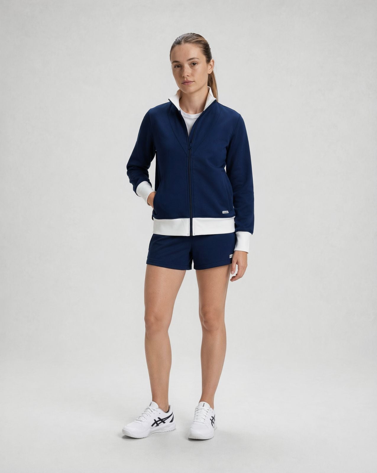 woman wearing navy jacket with white trim and matching navy shorts standing in studio, styled for padel and pickleball clothing