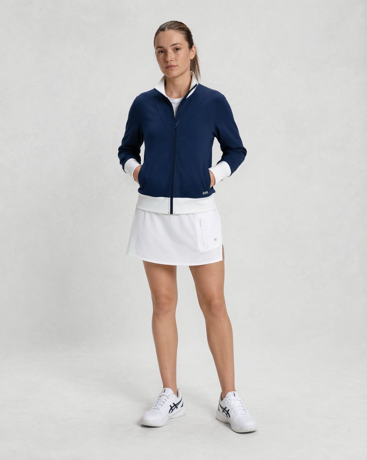 female model wearing navy zip-up jacket with white trim and white athletic skirt, styled for padel and pickleball apparel
